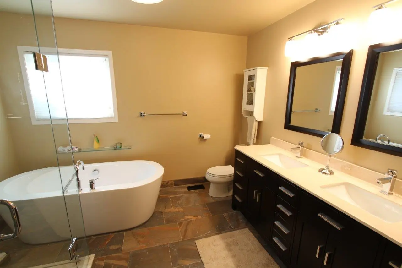 Modern bathroom with a freestanding white bathtub, double sink vanity with dark cabinets, two large mirrors, and a small wall-mounted cabinet above the toilet, featuring beige walls and a tiled floor.