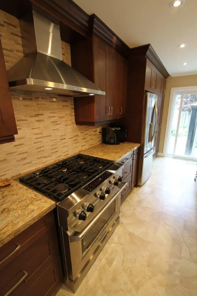 Modern kitchen with stainless steel gas stove and range hood, granite countertops, dark wooden cabinets, a coffee maker on the counter, stainless steel refrigerator, beige mosaic tile backsplash, and beige tiled floor leading to a sliding glass door.