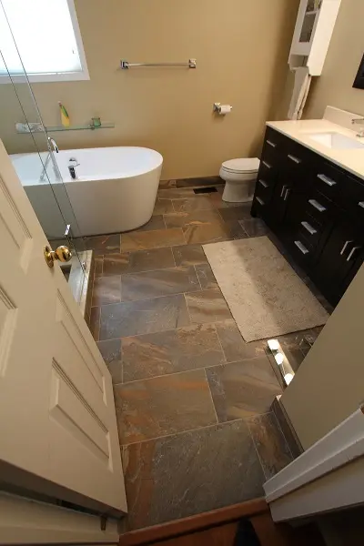 View of a modern bathroom with a freestanding white bathtub, toilet, dark wood vanity with white countertop, beige walls, tiled floor, and a beige bath mat near the vanity.