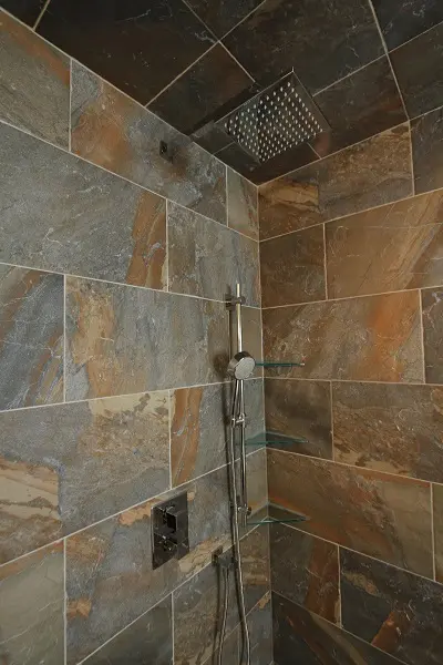Corner of a shower with brown and gray large stone tiles, featuring a square overhead rain showerhead, a handheld showerhead on a vertical rail, and three small glass corner shelves.