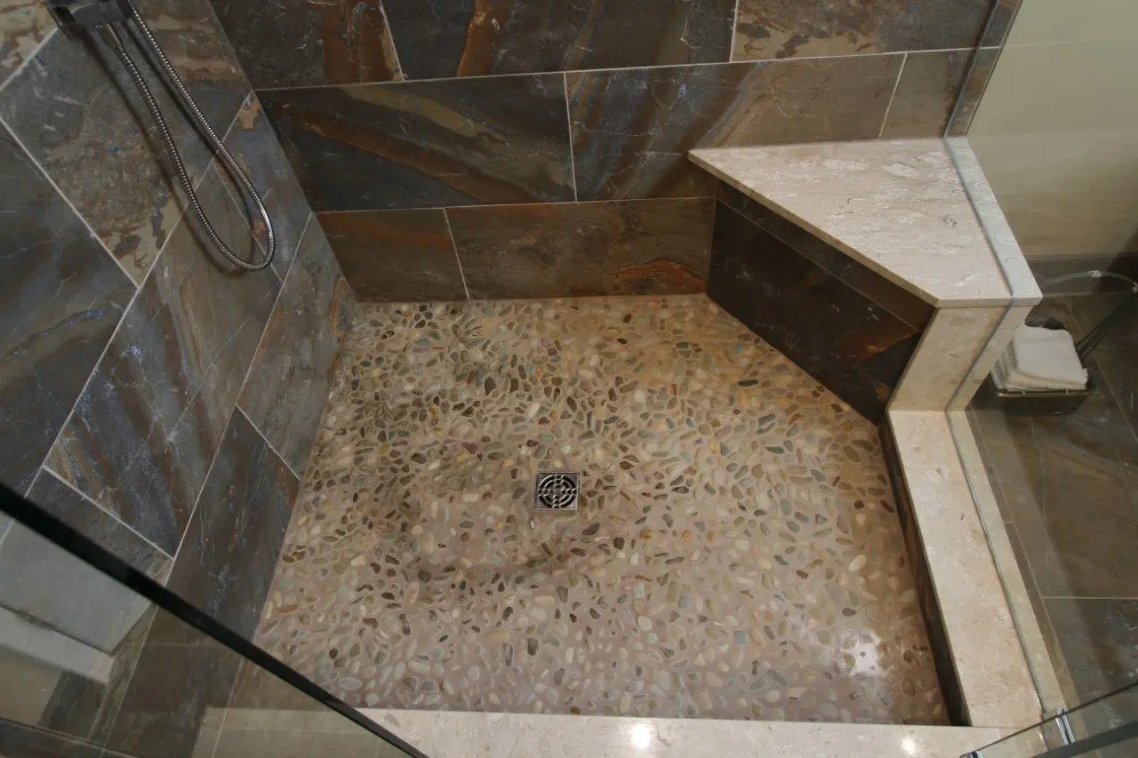 Shower area with pebble stone floor, dark brown and gray tiled walls, a corner bench with beige stone top, and a metal drain in the center.