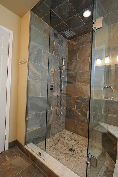 Modern walk-in shower with glass doors, stone tile walls and pebble floor, featuring a rainfall showerhead and a handheld shower wand.
