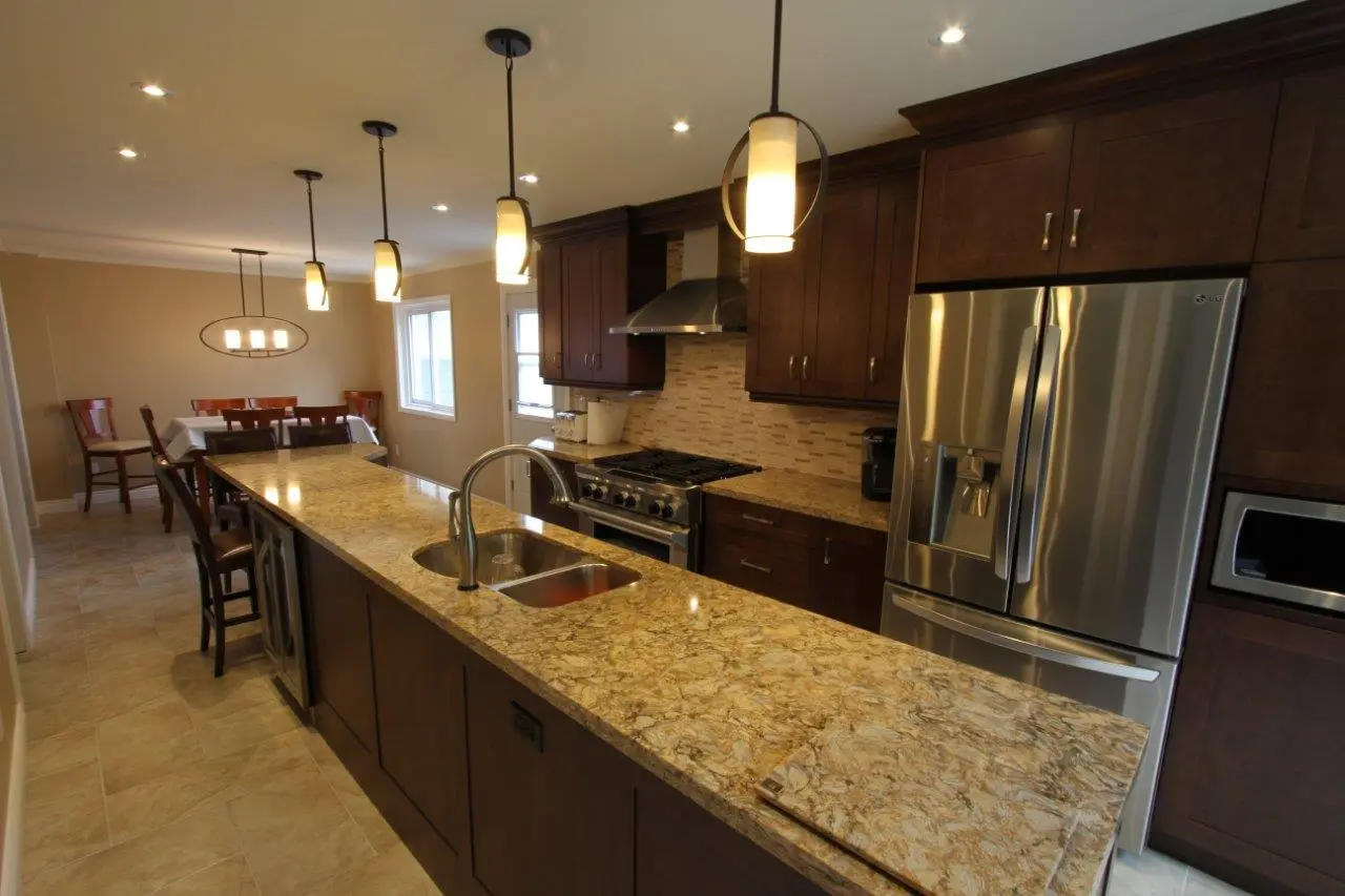 Modern kitchen with long granite countertop island featuring a stainless steel double sink and multiple pendant lights above, dark wood cabinetry, a stainless steel refrigerator and oven, and a dining area with chairs and table in the background.