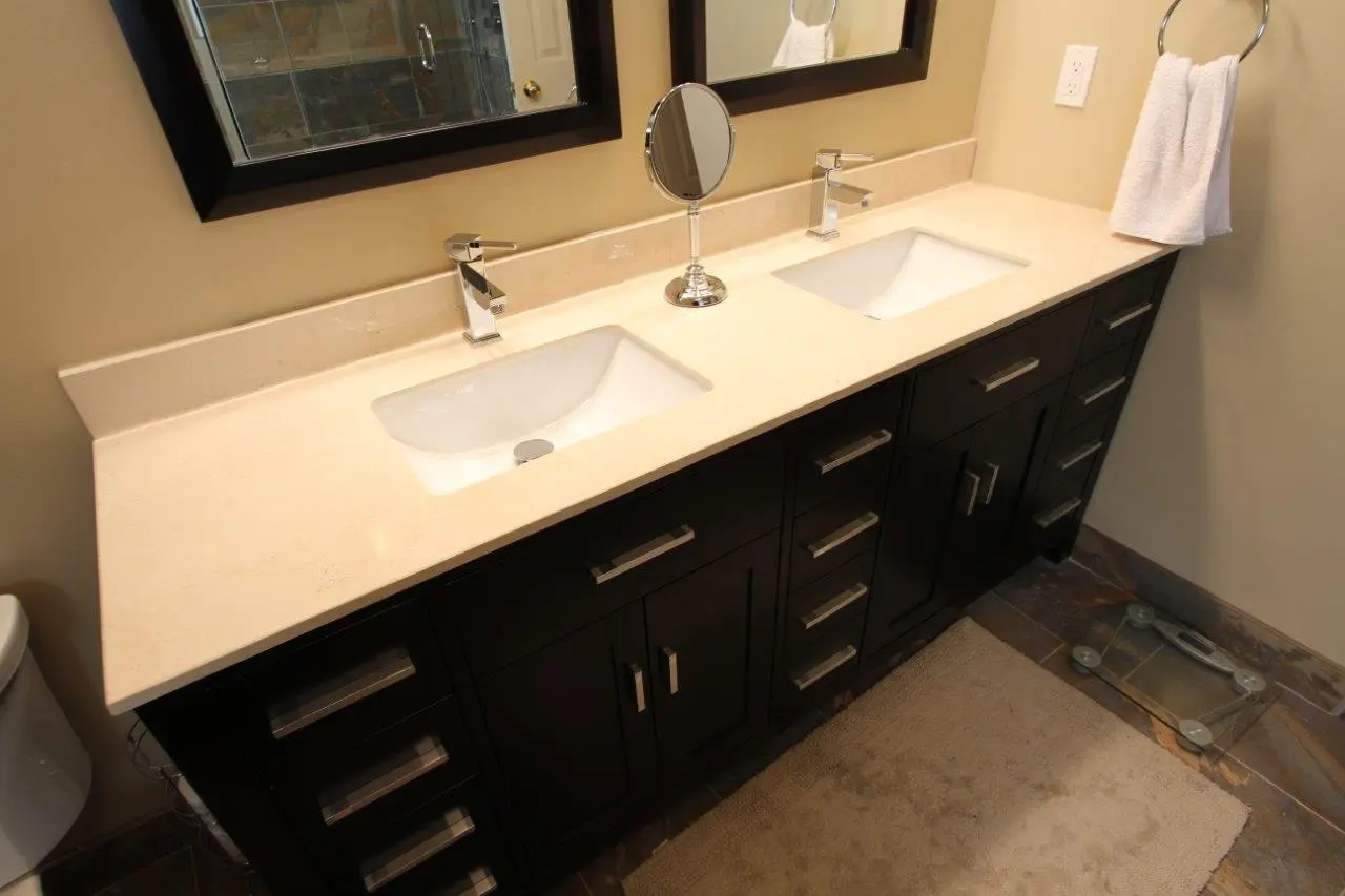 Bathroom vanity with a cream-colored countertop, two white rectangular sinks with chrome faucets, a round tabletop mirror in the center, dark wood cabinets with silver handles underneath, a beige wall with a towel hanging on a chrome ring, a portion of a white toilet visible on the left, and a beige bath mat on the floor next to a glass scale.