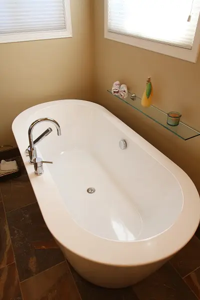 White freestanding bathtub with modern faucet in a corner bathroom with beige walls, glass shelf holding rolled towels, a yellow decorative item, and a candle, and two windows with blinds.