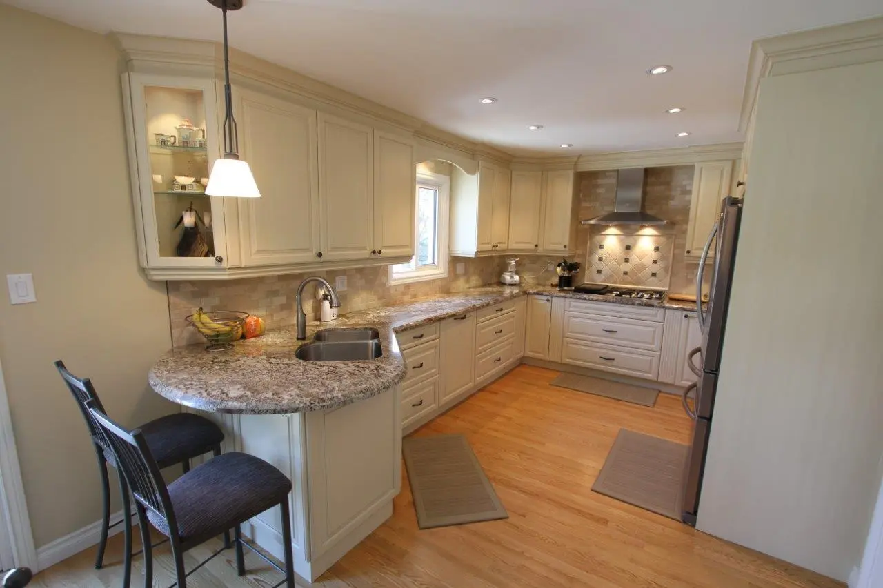 Modern kitchen with beige cabinets, granite countertops, a double sink, stainless steel refrigerator and stove, wooden flooring, two black bar stools, and three floor mats, illuminated by pendant and recessed lighting.