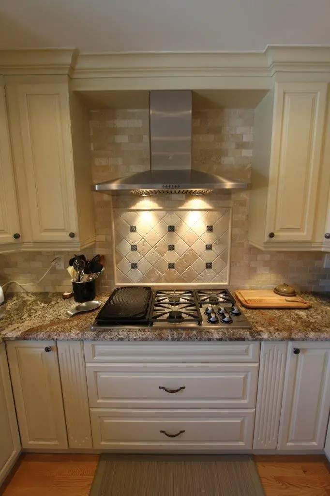 Modern kitchen stove area with a stainless steel range hood, beige tiled backsplash with decorative square tiles, granite countertop, utensil holder with black and silver utensils, and a wooden cutting board.