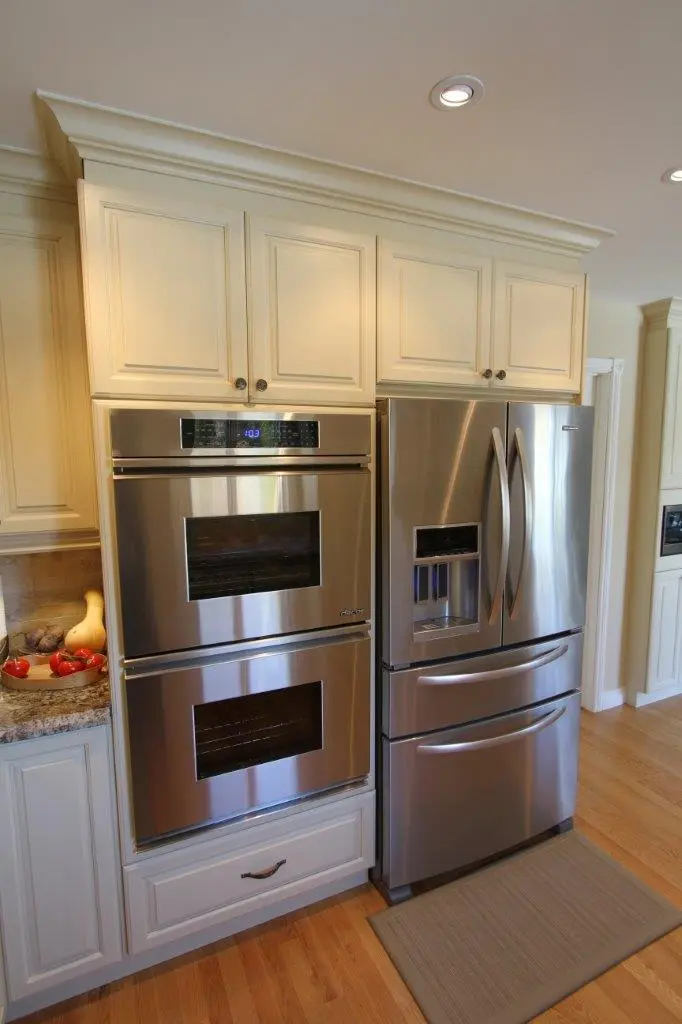 Modern kitchen with stainless steel double wall oven and French door refrigerator with two freezer drawers, surrounded by cream-colored cabinetry and hardwood floor.
