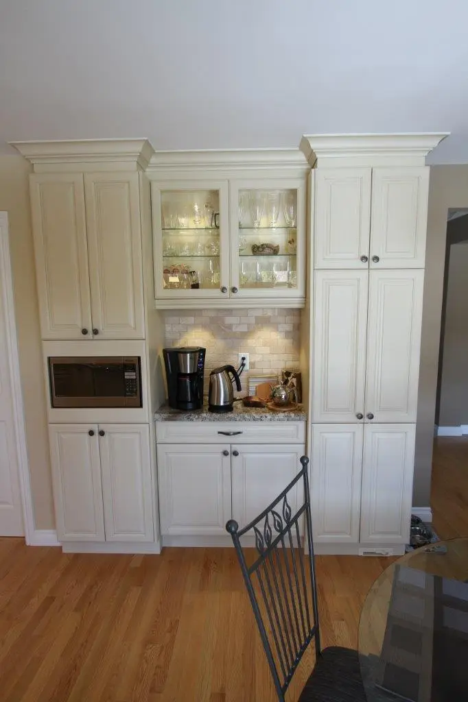 Built-in kitchen cabinetry with cream-colored cabinets, a microwave, glass display cabinets with glassware, and a countertop holding a coffee maker, electric kettle, and other kitchen items, with hardwood flooring and a black metal chair with vertical and decorative backrest in the foreground.