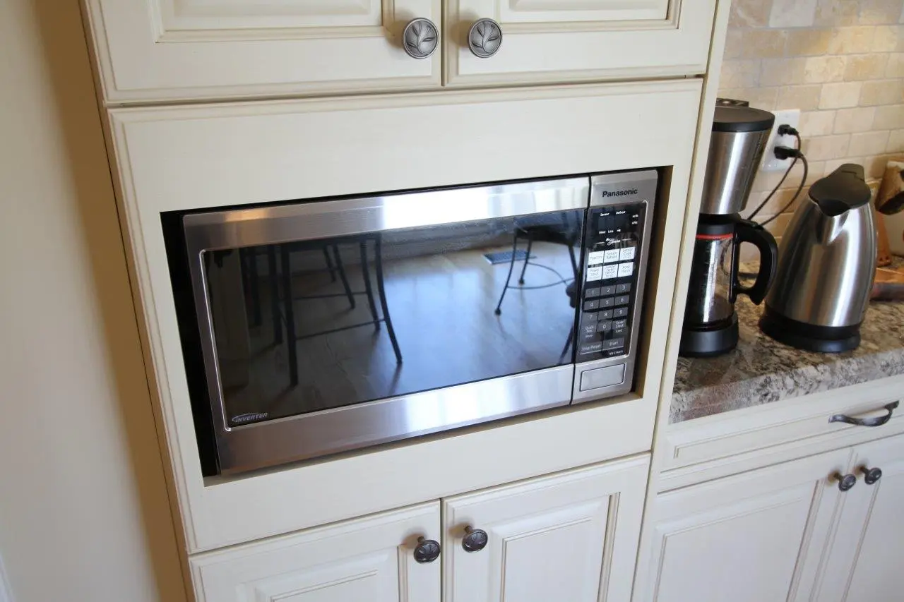 Built-in stainless steel Panasonic microwave oven installed in white kitchen cabinetry with a granite countertop partially visible to the right, featuring a coffee maker and electric kettle.