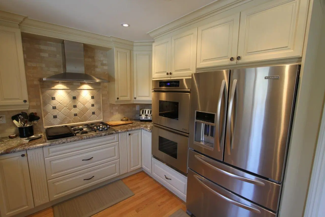 Modern kitchen corner with cream-colored cabinets, stainless steel double oven, French door refrigerator with water dispenser, gas stovetop with overhead stainless steel hood, toaster, and cooking utensils on granite countertops, hardwood floor with beige mats.