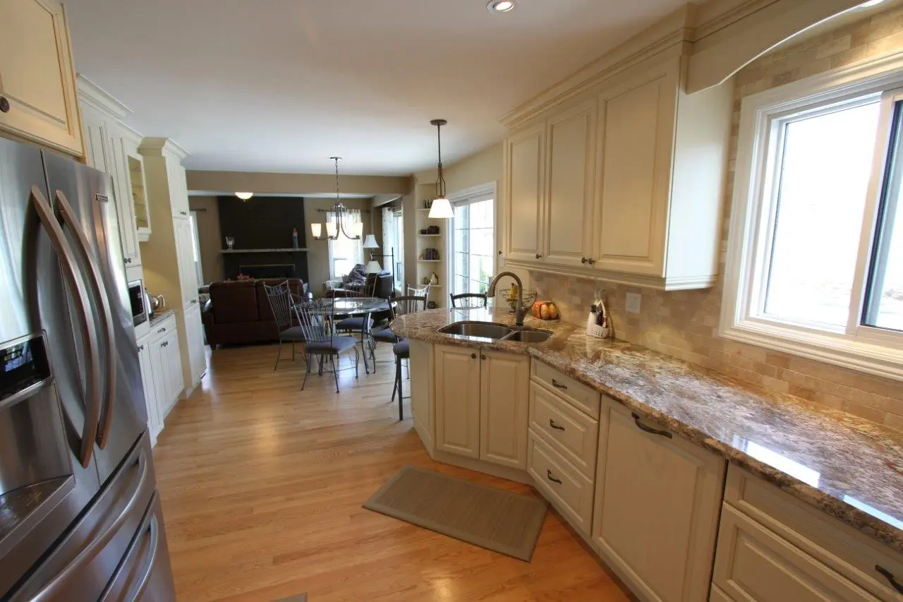 Modern kitchen with beige cabinets, granite countertops, stainless steel refrigerator, and a double sink, opening into a dining area with a round glass table, metal chairs, and a living room with brown sofas and a fireplace.