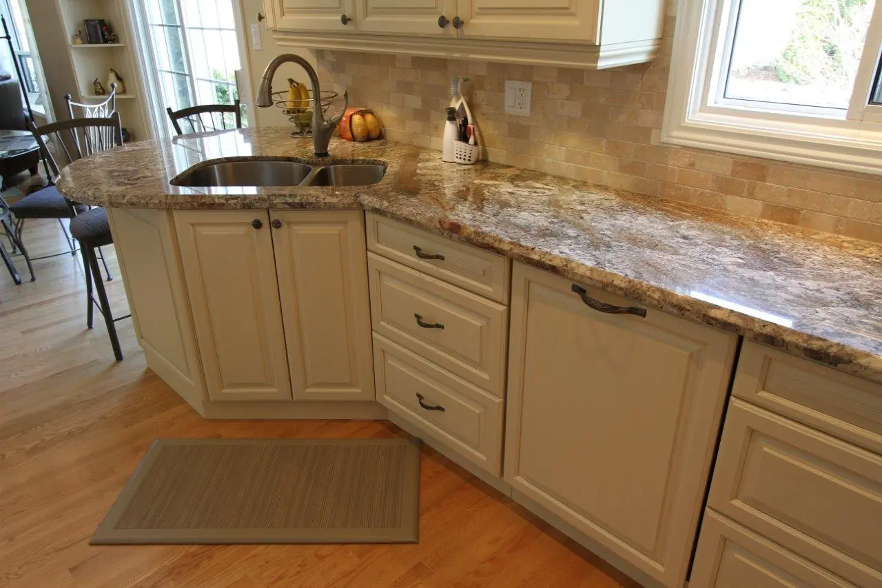 Kitchen countertop with a dual stainless steel sink, beige cabinets and drawers, a brown cushioned mat on light wood flooring, and a window above the counter allowing natural light in.