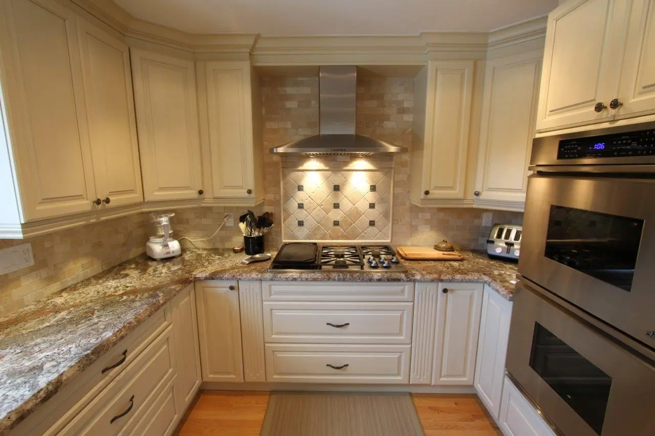 Modern kitchen corner with beige cabinets, granite countertops, stainless steel oven and stove with vent hood, a toaster, blender, cutting board, and kitchen utensils holder on the counter, with warm lighting illuminating the stove area and hardwood floor.