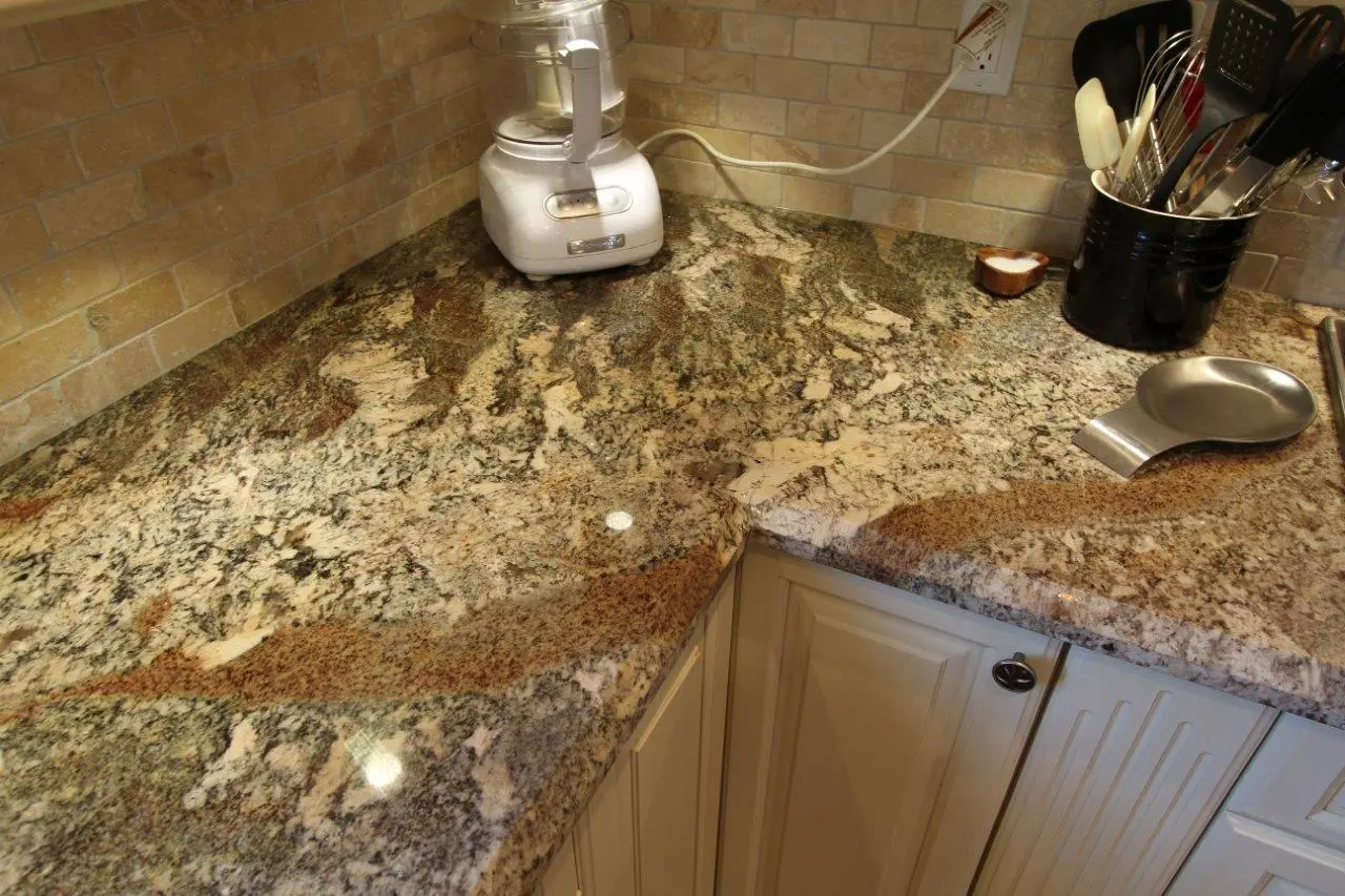 Granite kitchen countertop with beige, brown, and gray patterns, featuring a white food processor, a black container holding kitchen utensils, and a metal spoon rest, set against a beige tiled backsplash and cream-colored cabinets.