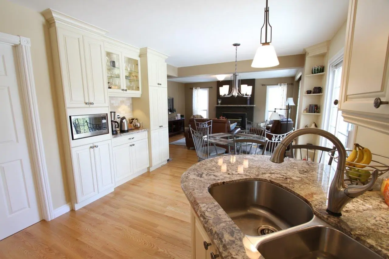 Open kitchen with granite countertop, double stainless steel sink, and light cream cabinetry with built-in microwave, leading to a dining area with glass table and metal chairs, and a living room with brown leather sofas and a fireplace.