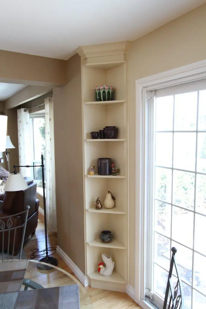 Narrow cream-colored corner shelf unit in a living room, displaying various pottery items including vases, pitchers, and decorative figurines, next to a large window with white trim and a view of the outdoors.