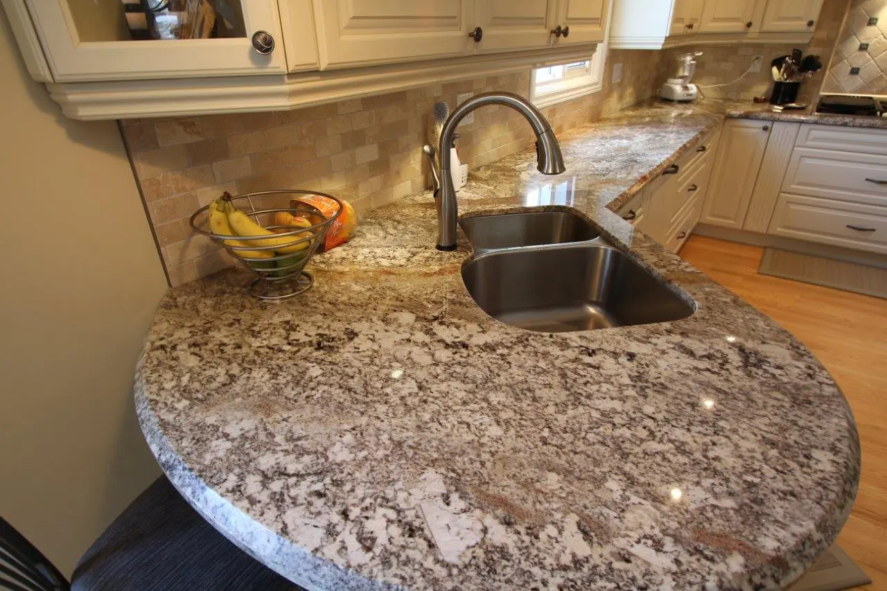 Polished granite kitchen countertop with a double stainless steel sink and a curved edge, featuring a metal fruit basket holding bananas and oranges, beige tiled backsplash, white cabinets, and wooden flooring.