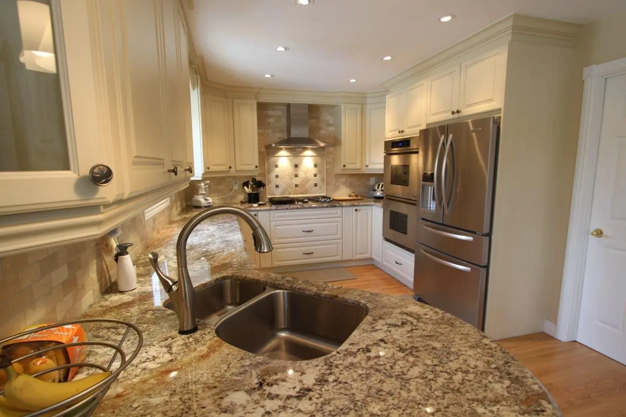 Modern kitchen with beige granite countertops, a double stainless steel sink with a faucet, cream-colored cabinets, stainless steel oven and refrigerator, wooden floor, and a bowl of fruit on the counter.