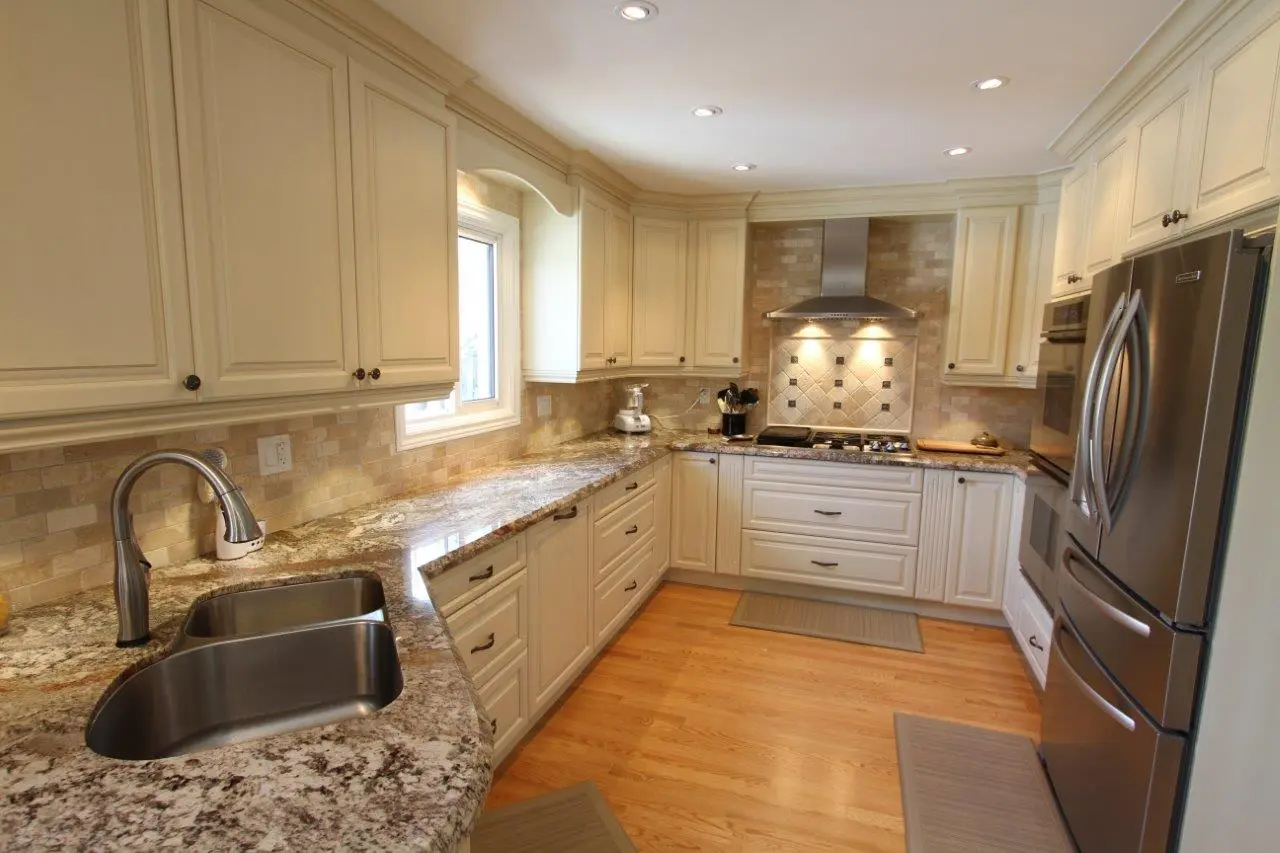 Modern kitchen with cream-colored cabinets, granite countertops, stainless steel double sink, stainless steel appliances including a refrigerator and stovetop with range hood, beige tiled backsplash, and hardwood flooring with gray floor mats.