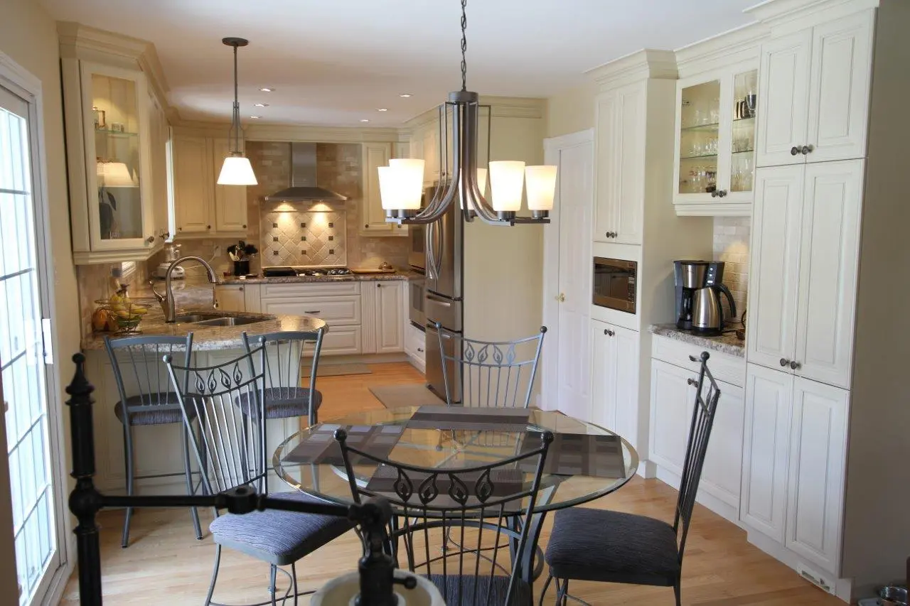 Bright kitchen with cream-colored cabinets, granite countertops, stainless steel appliances, a glass round dining table with black metal chairs, pendant and chandelier lights, and a coffee maker on the counter.