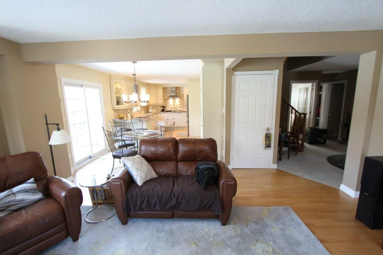 Living room with light brown leather armchairs and sofa, a round glass-top dining table with metal chairs near a bright window, beige walls, wood flooring, a white door, and a view into a kitchen with white cabinetry and a tiled backsplash.