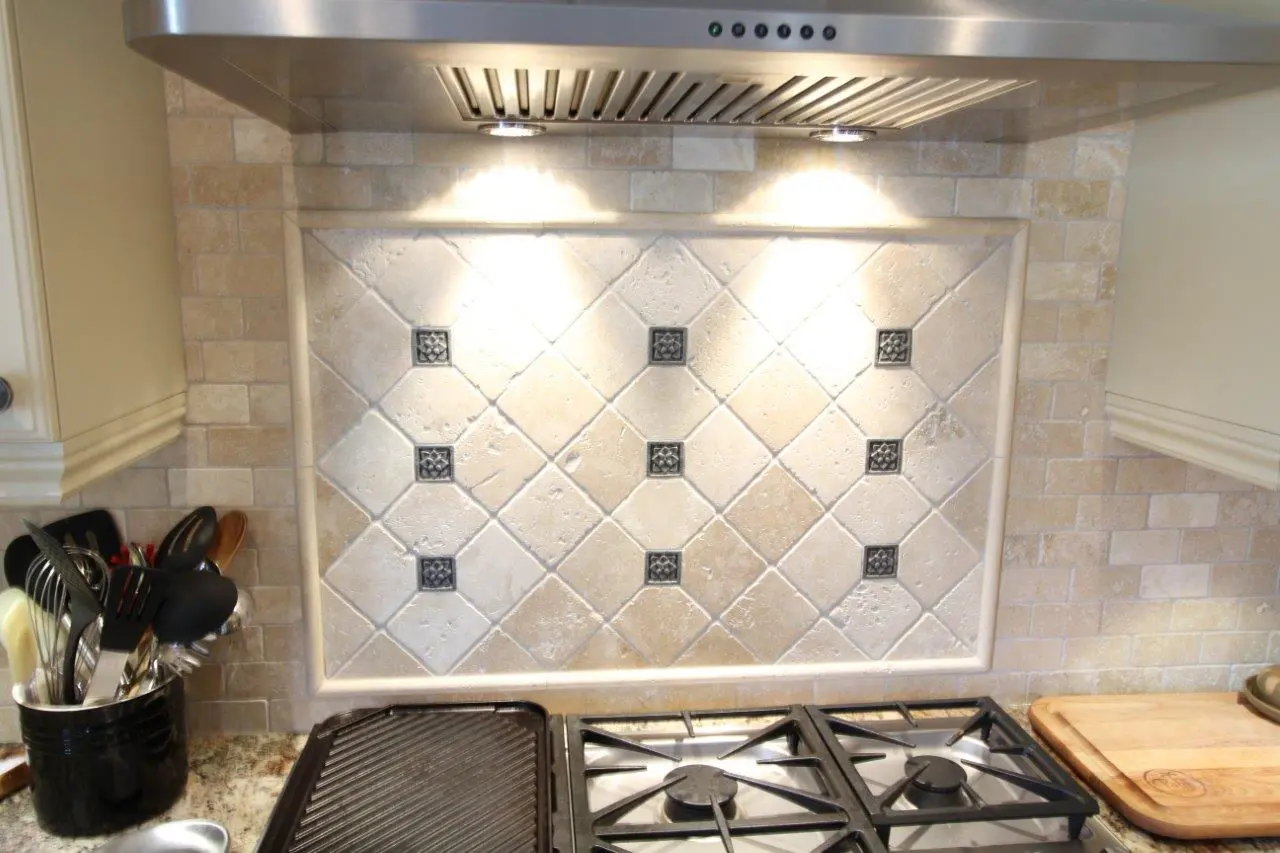 Kitchen stove area with a stainless steel range hood and tiled backsplash featuring beige and white tiles with decorative black square accents, a black utensil holder with various cooking tools on the left, and a wooden cutting board on the right.