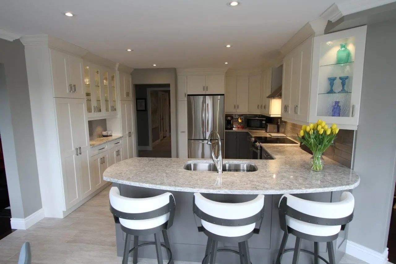 Modern kitchen with white cabinetry, a large marble countertop island with a built-in sink and three black and white bar stools, stainless steel appliances, glass cabinet displaying blue vases, and a vase of yellow tulips on the countertop.