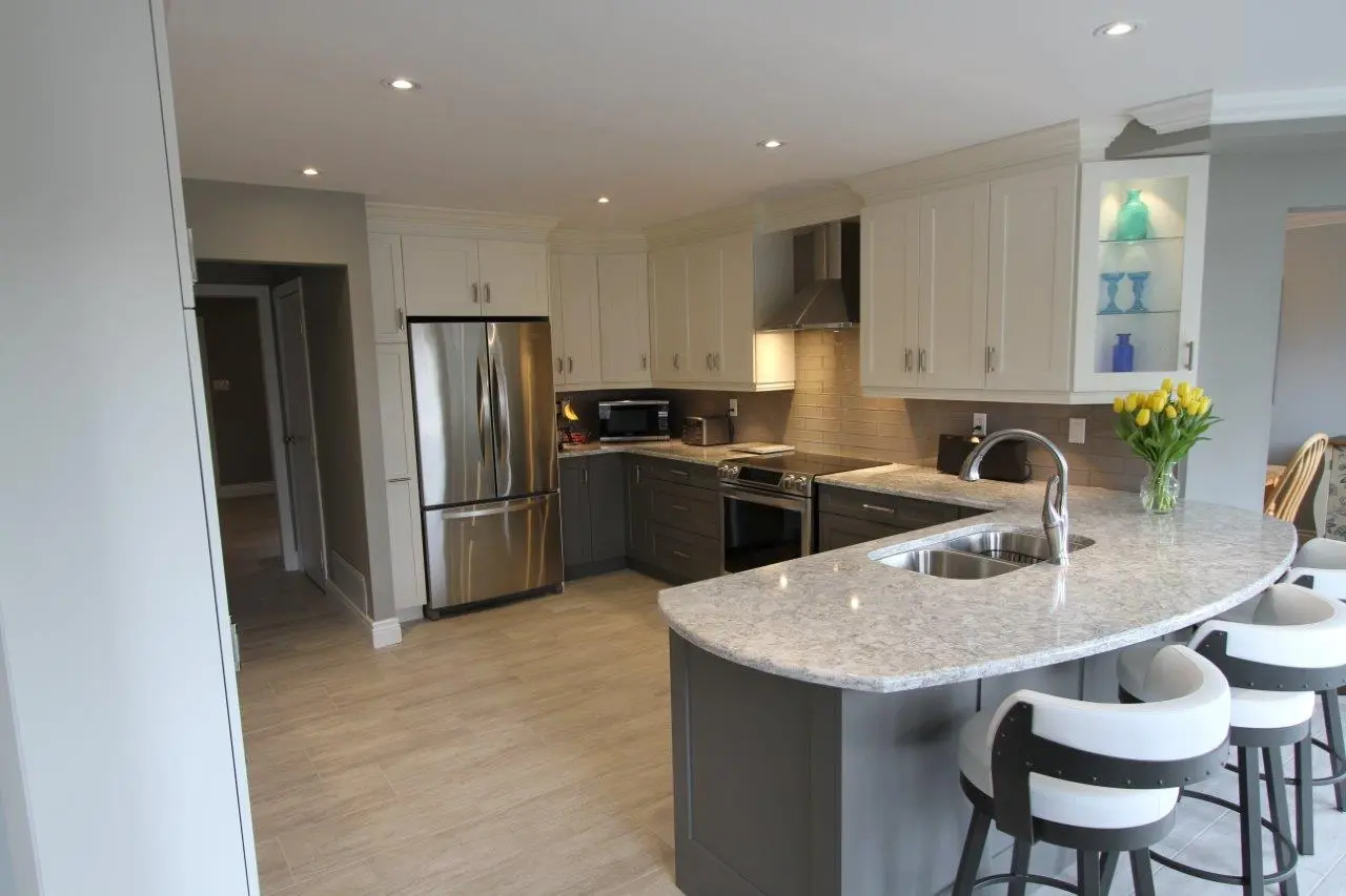 Modern kitchen with a curved marble countertop island featuring a sink and three white and black bar stools, white upper cabinets, gray lower cabinets, stainless steel refrigerator and oven, glass shelves with decorative blue bottles, gray backsplash, and a vase of yellow tulips on the countertop.
