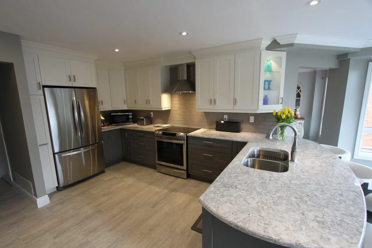Modern kitchen with a curved marble countertop, dual sinks, stainless steel refrigerator and stove, white upper cabinets, dark lower cabinets, and a vase of yellow tulips on the counter.