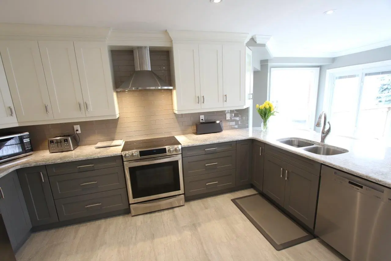 Modern kitchen with gray lower cabinets and white upper cabinets, stainless steel appliances including an oven with a range hood, a dishwasher, a toaster, and a microwave, granite countertops, a double sink under large windows, and a vase with yellow flowers on the countertop.