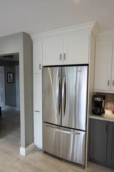 Stainless steel French door refrigerator with bottom freezer drawer integrated into white kitchen cabinetry next to a coffee maker on a gray cabinet with a granite countertop.