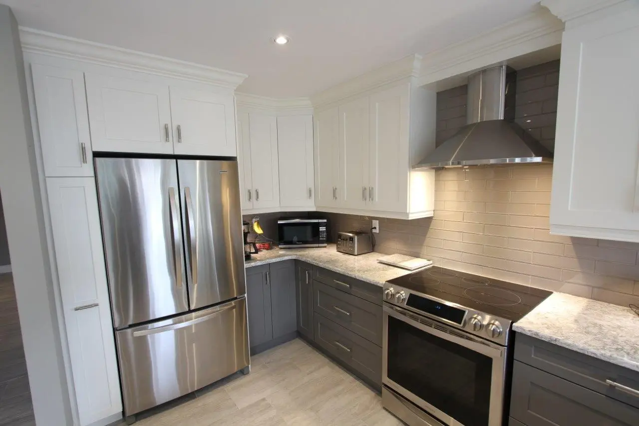 Modern kitchen corner with stainless steel refrigerator, stove with range hood, and microwave, featuring white upper cabinets, gray lower cabinets, and light-colored countertops and tiled backsplash.