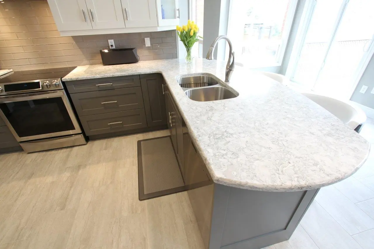Modern kitchen with gray cabinets, a light gray marble countertop island featuring a double stainless steel sink and curved edge, stainless steel oven, beige tile backsplash, light wood flooring, and a vase of yellow tulips near a large window providing natural light.