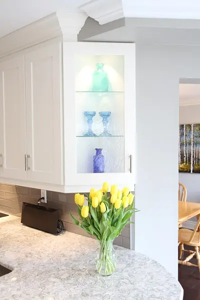 Yellow tulips in a clear glass vase on a kitchen countertop with white cabinets and a glass-door cabinet displaying blue and green glassware, with a dining room visible in the background