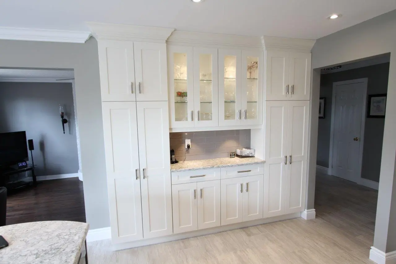 White built-in kitchen cabinetry with glass display doors on top, a granite countertop, and gray tile backsplash, situated between two doorways in a modern kitchen with light gray walls and light-colored floor tiles.