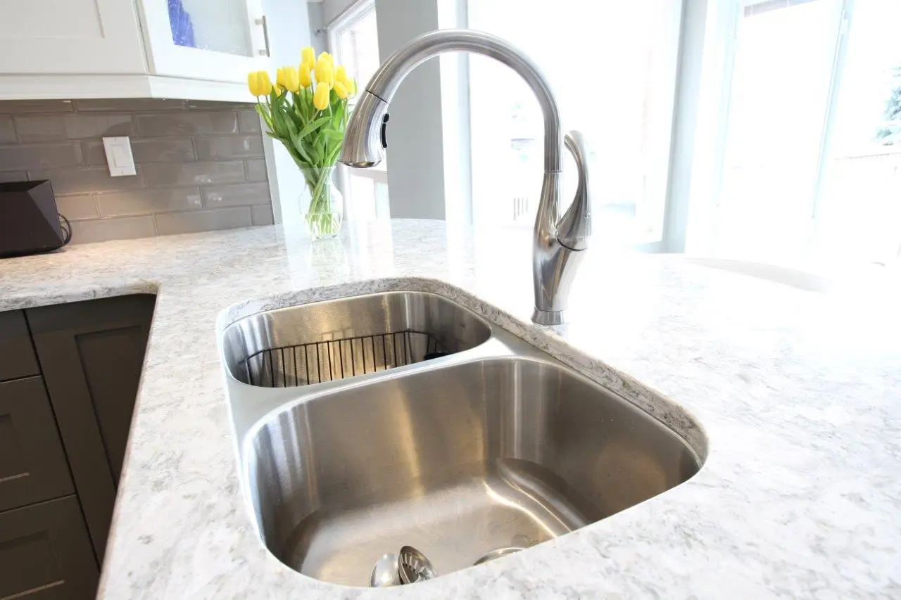 Modern kitchen sink with a dual-basin stainless steel design set in a light-colored marble countertop, with a curved silver faucet and a vase of yellow tulips in the background near a window.