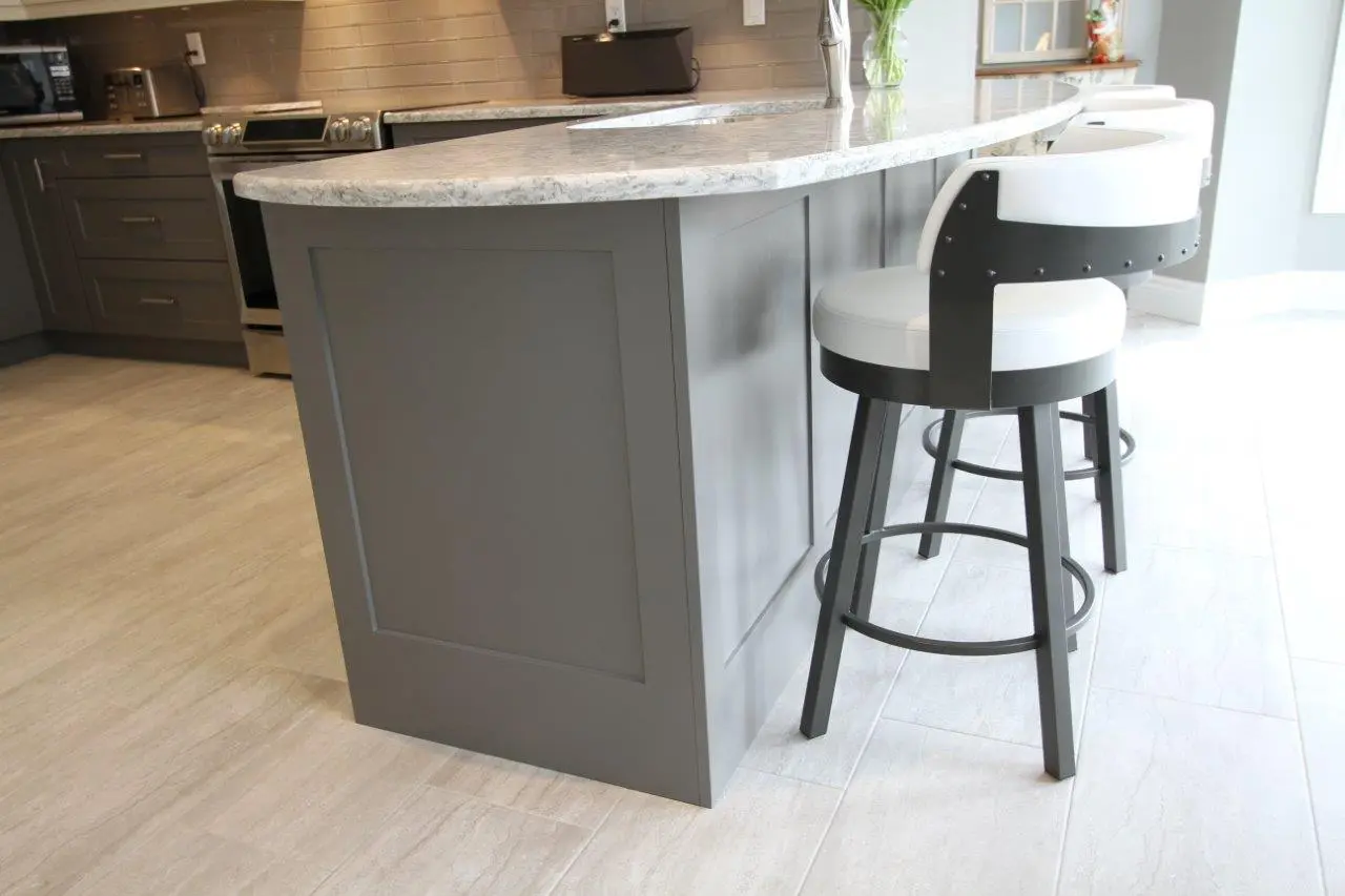 Modern kitchen island with a gray base and a light granite countertop, accompanied by white cushioned bar stools with black frames on a light tile floor.