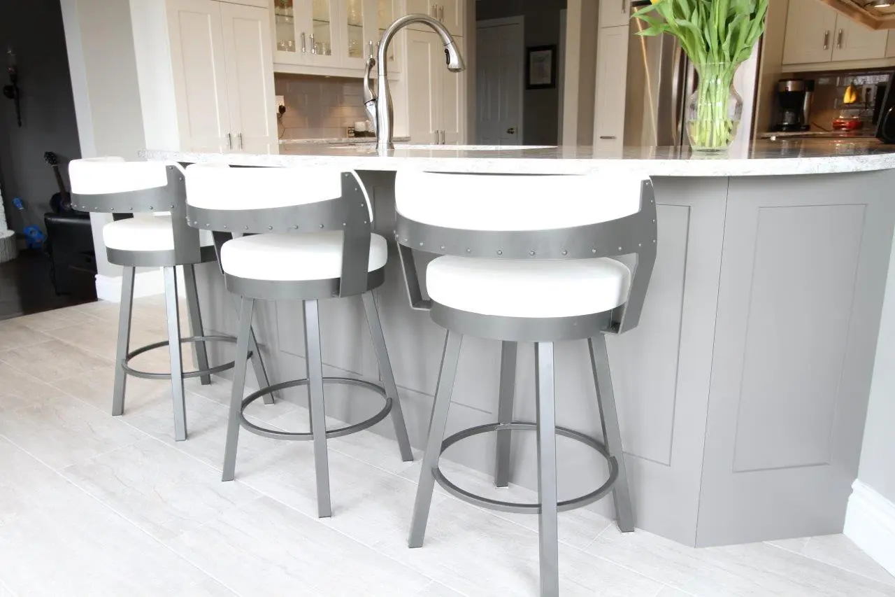 Three modern bar stools with white cushioned seats and backs and gray metal frames lined up along a gray kitchen island with a white marble countertop and a tall silver faucet, with a vase of green tulips on the counter, and white tiled floor below.