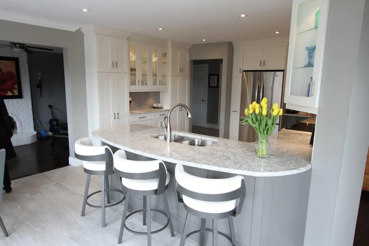 Modern kitchen with a curved marble countertop island featuring a dual sink and a vase of yellow tulips, three gray and white swivel bar stools, white cabinetry, stainless steel refrigerator, and recessed ceiling lights.
