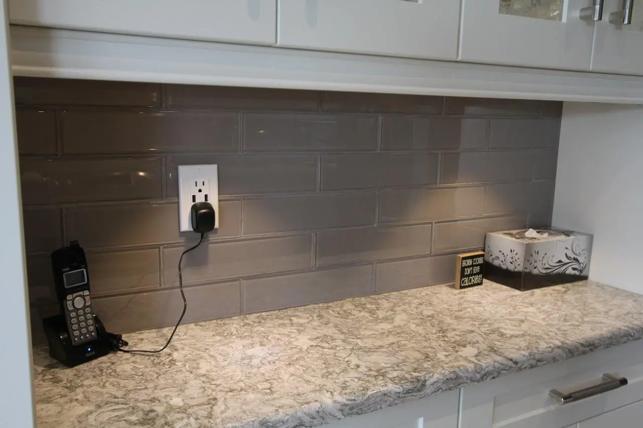 Marble countertop with a gray subway tile backsplash, a cordless phone charging on the left, and a decorative tissue box with a small sign that says 'Brown cookies don’t have calories!' on the right under white cabinets.