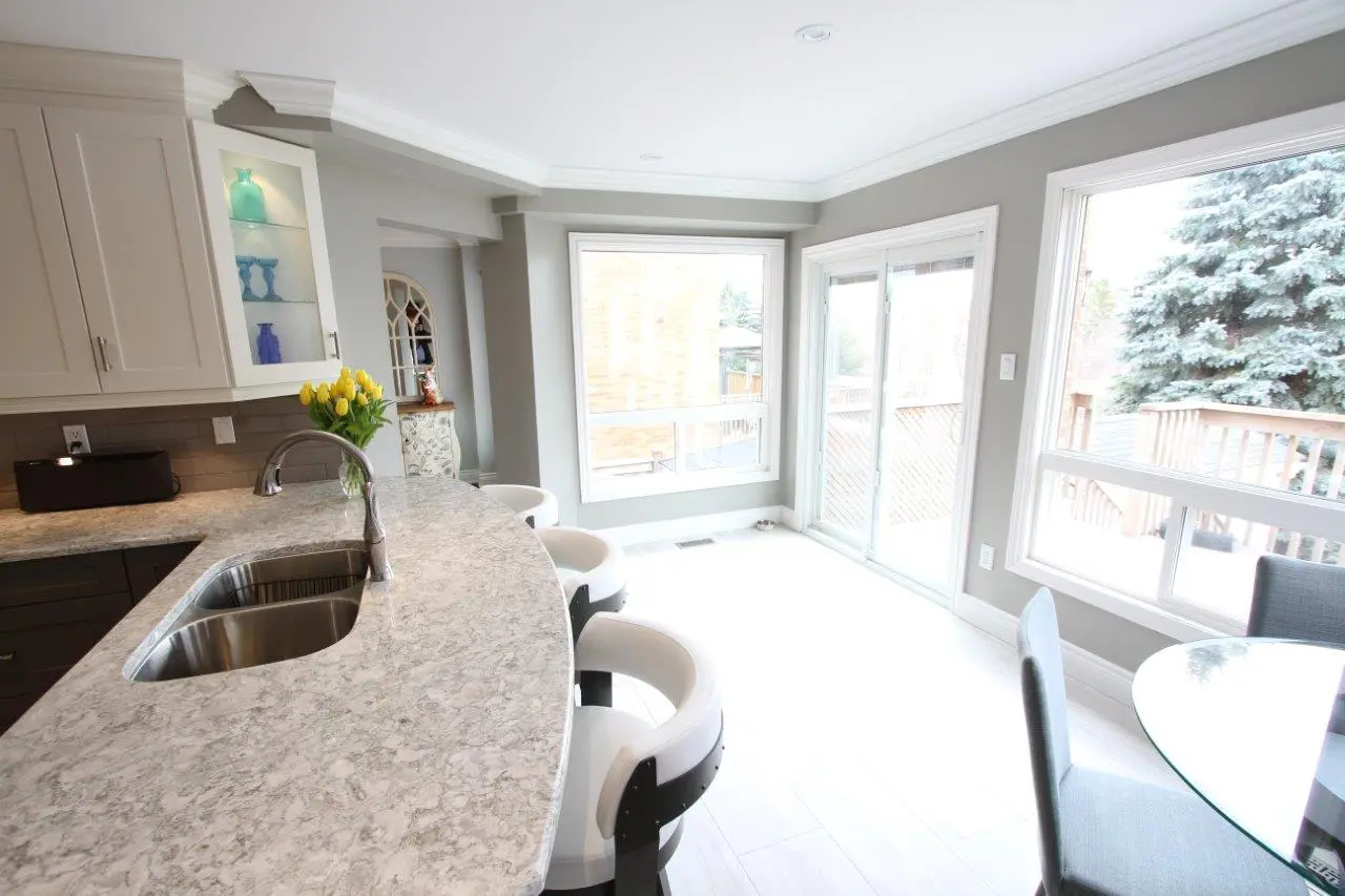Bright modern kitchen with a marble countertop island featuring a double sink and chrome faucet, white cushioned bar stools, light gray cabinets with a glass display, and large windows and sliding glass doors letting in natural light revealing an outdoor deck with trees.