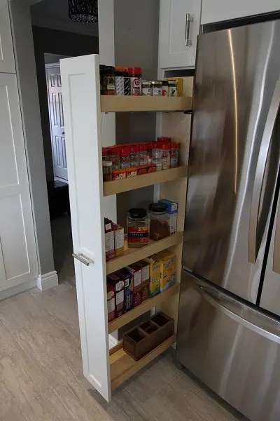 Narrow pull-out pantry next to a stainless steel refrigerator containing shelves stocked with spice bottles, small containers, and boxed food items in a modern kitchen with light wood flooring and white cabinetry.