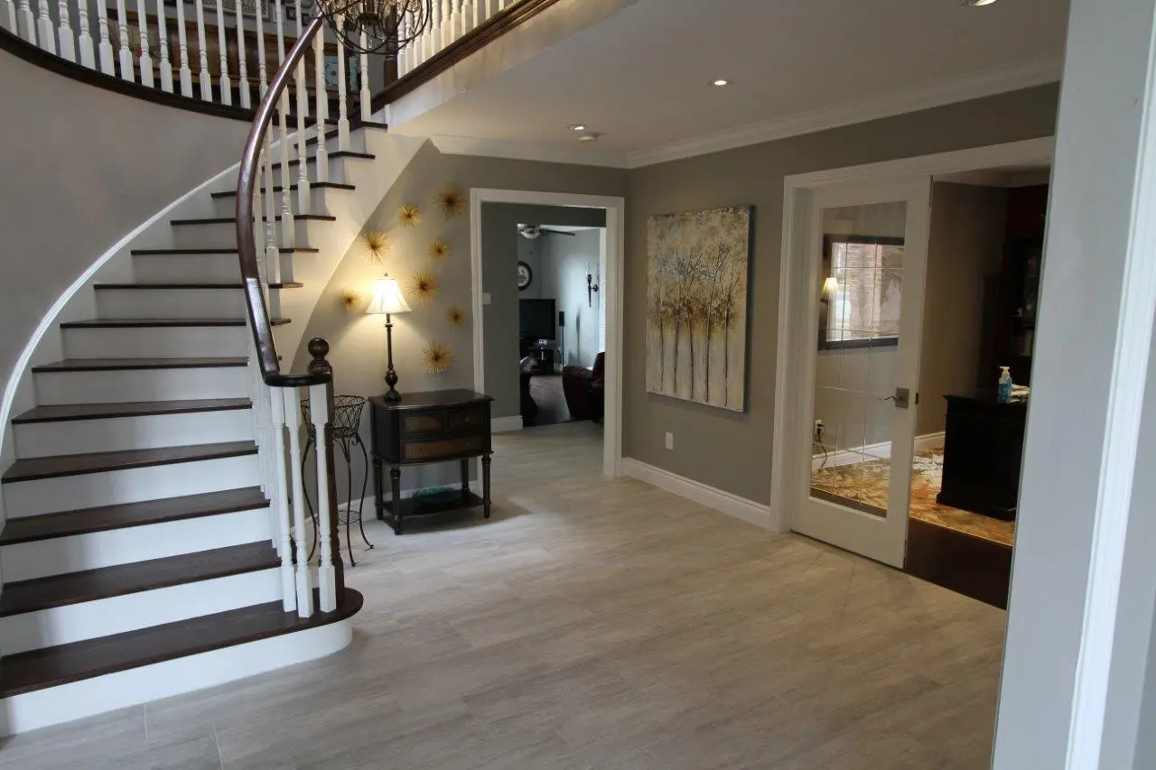 Interior of a modern home foyer with a curved wooden staircase on the left, light gray walls, a small dark wooden table with a lamp and decorative gold wall art, and two doorways leading to other rooms, one with a glass-paned door and a painting of trees on the wall.
