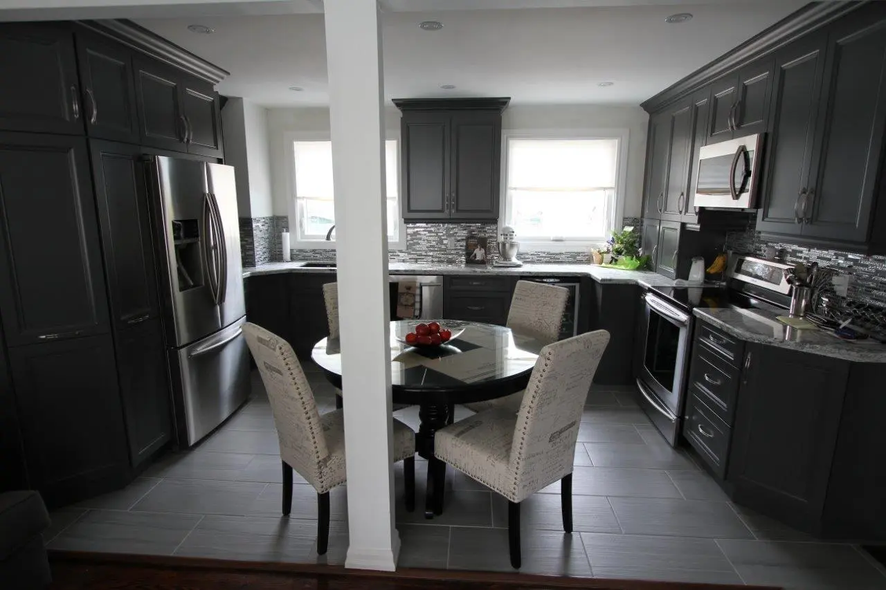 Modern kitchen with dark gray cabinets and stainless steel appliances, a round black glass dining table with a bowl of red tomatoes, surrounded by four beige upholstered chairs with printed text, and a white column in the center.