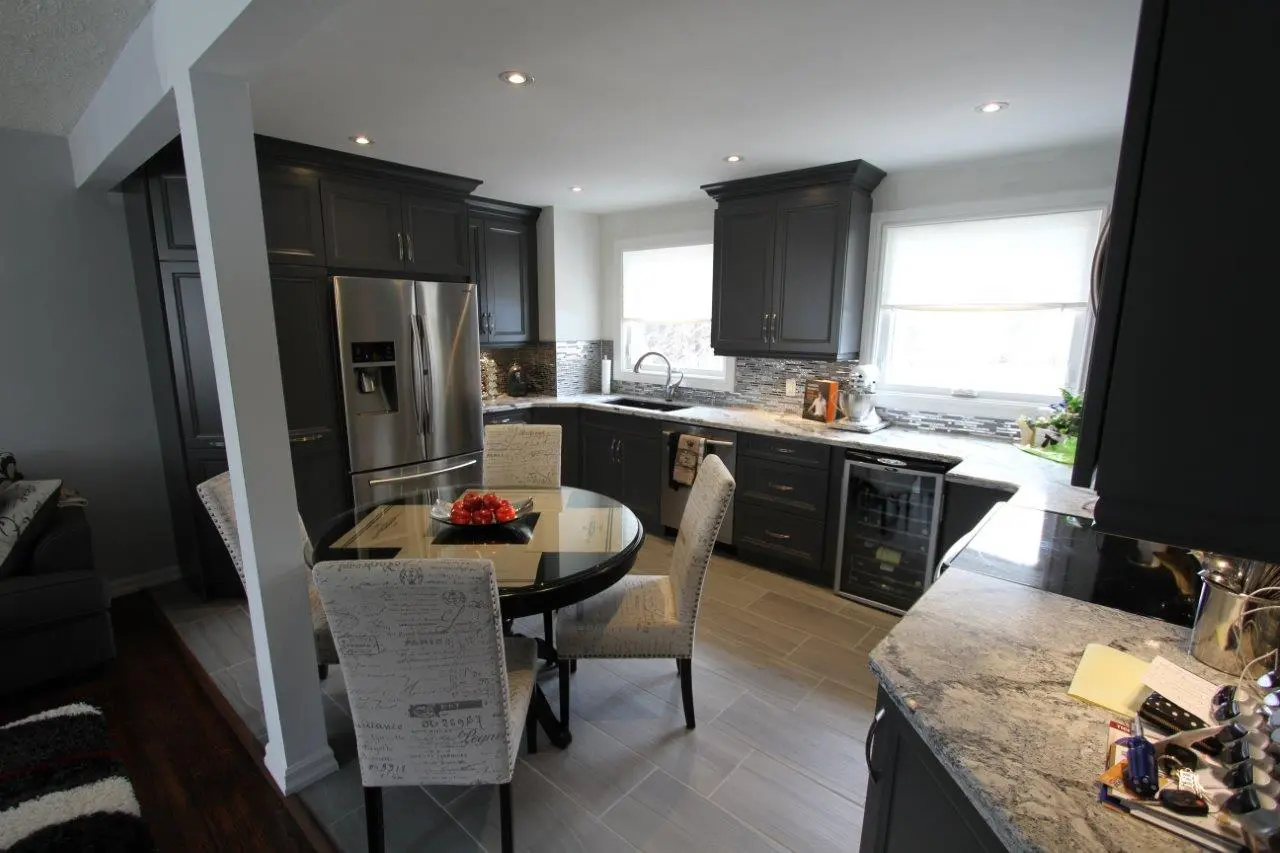 Modern kitchen with dark gray cabinets, stainless steel refrigerator, and marble countertops, featuring a round glass-top dining table with four beige upholstered chairs and a centerpiece of red apples.