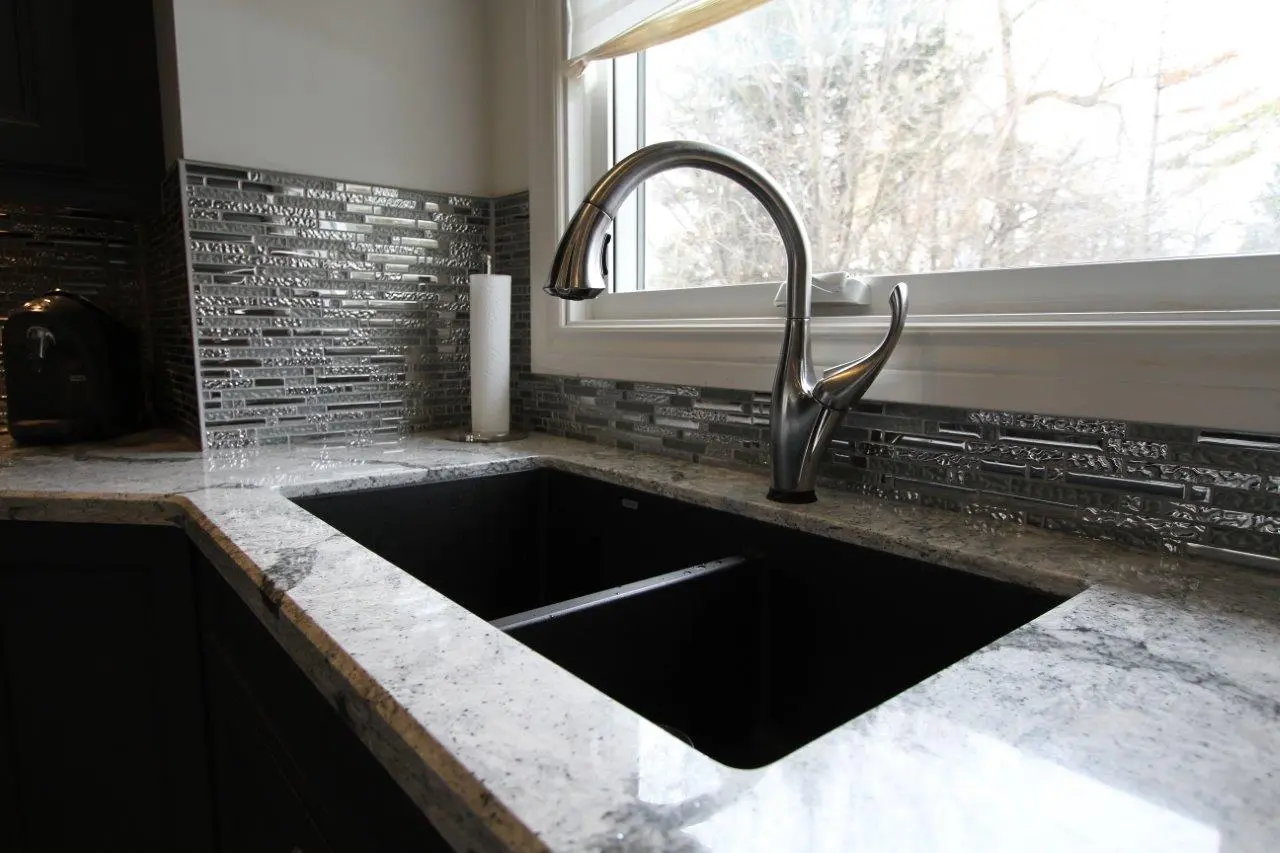 Modern kitchen sink with a dual black basin, silver curved faucet, granite countertop, and decorative metallic tile backsplash, positioned under a window with an outside view of trees.