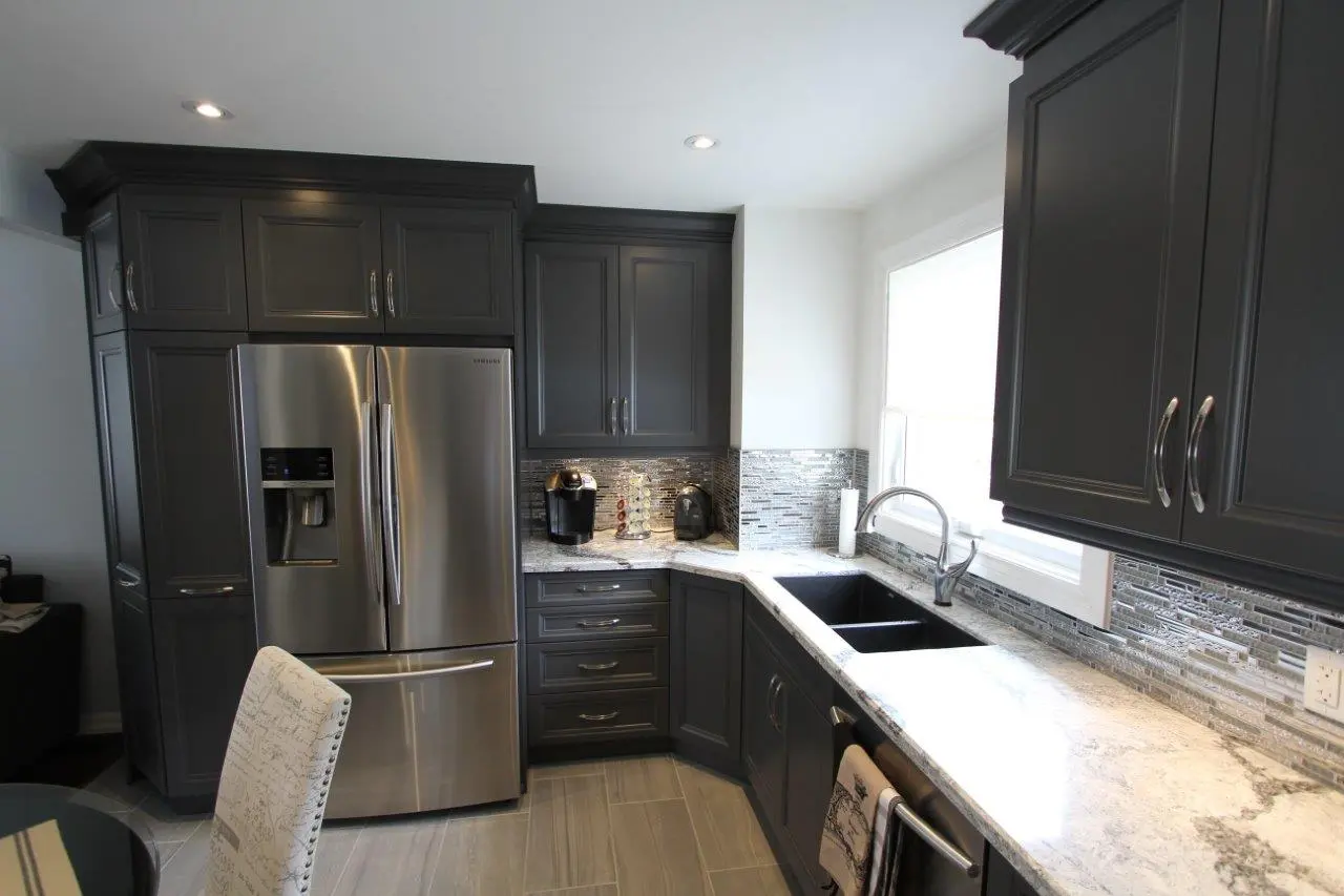 Modern kitchen with dark gray cabinets, stainless steel refrigerator, marble countertop, black double sink, mosaic tile backsplash, coffee maker, and light gray tiled floor.