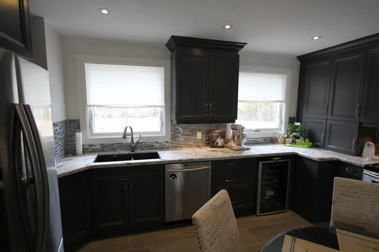 Modern kitchen with dark cabinets, marble countertops, two windows with white blinds, stainless steel sink and dishwasher, wine cooler, and floral decorations on the corner counter.