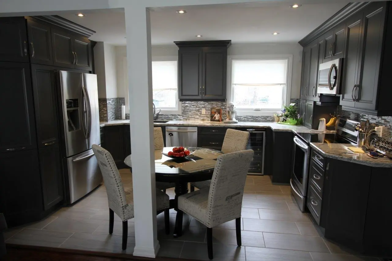 Modern kitchen with dark gray cabinets, stainless steel refrigerator, oven, microwave, and dishwasher, a round glass table with four beige upholstered chairs, and two large windows providing natural light.